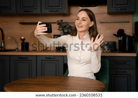A young brunette woman makes a video call from her smartphone while sitting in the kitchen at home. 
