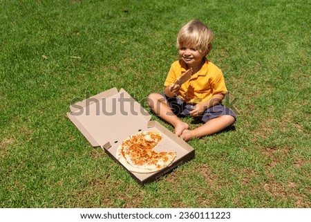 little blond boy eats pizza in the park sitting on the grass