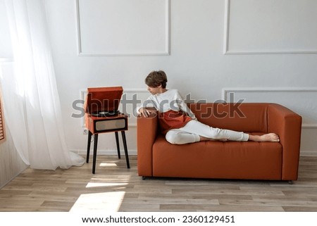 Teenager boy sitting on brown sofa and looking on brown retro wooden turntable with vinyl record. Nostalgic music listening, lifestyle, home interior. Royalty-Free Stock Photo #2360129451