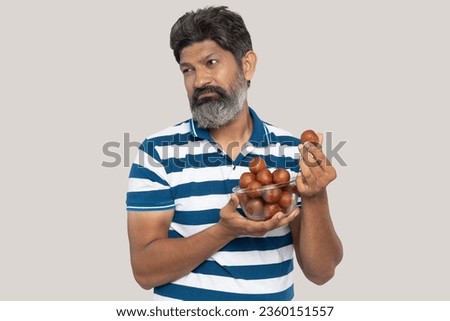 Portrait of a Indian beard man showing unhappy facial expressions to see a Gulab jamub. eating sweet Gulab Jamun against white studio background Royalty-Free Stock Photo #2360151557