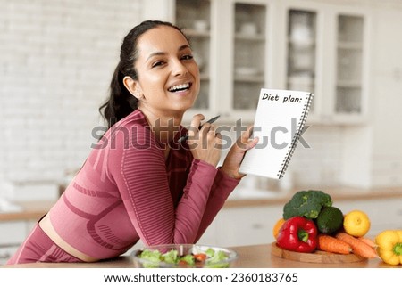Sports Nutrition. Happy Fit Young Lady Writing Her Diet Plan In Notebook, Planning Recipes And Meals For Weight Loss Posing Near Table In Modern Kitchen Interior At Home, Smiling To Camera