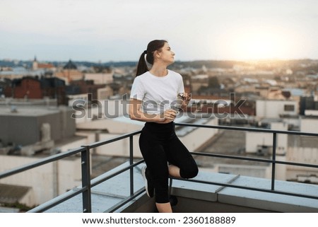 Relaxed health lady in activewear watching city view while leaning on handrails in corner of building roof. Charming yogini with water bottle staring at evening sky after finishing outdoor session. Royalty-Free Stock Photo #2360188889