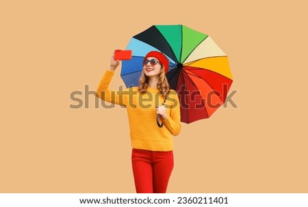 Autumn color style outfit, beautiful happy cheerful young woman holds colorful umbrella taking selfie with mobile phone wearing red french beret hat, yellow sweater on beige studio background
