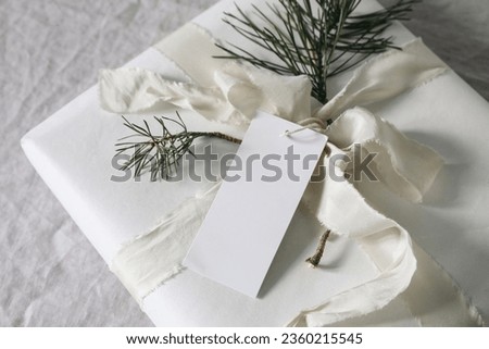 Beautiful Christmas present. Blank gift tag, label mockup. Handmade gift wrapping with cotton paper, pine tree branches and silk ribbon on linen table cloth.Winter birthday. Blurred background, top.