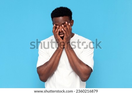 tired african american man in white t-shirt in depression covers his face with his hands on blue isolated background, sleepy indifferent man is bored and unhappy