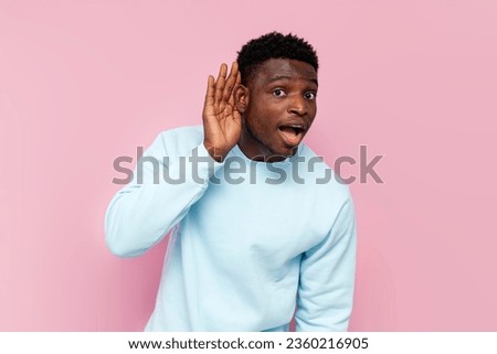 curious african american man in blue sweater eavesdrops on pink isolated background, the guy holds his hand near his ear and listens Royalty-Free Stock Photo #2360216905
