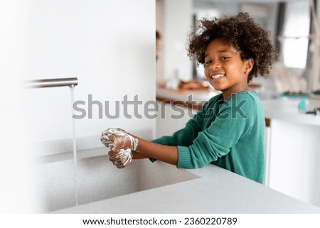 Happy young African American girl washing hands with soap smiling and looking at camera while standing at kitchen sink.