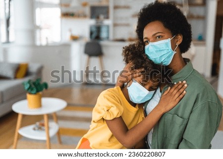 Sick and afraid African American girl resting head on mother's shoulder. Caring single mother hugging and consoling her young child while wearing protective masks..