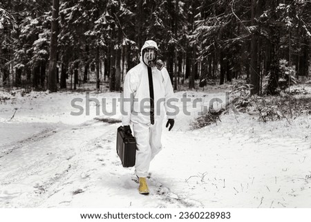 Scientist dosimetrist with radiometer and scientist radiation supervisor in protective clothing and gas masks examine a sample of radioactive material in hazardous radioactive zone. Royalty-Free Stock Photo #2360228983