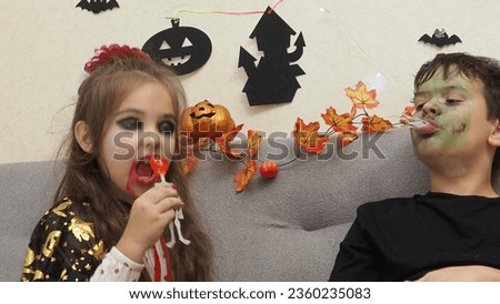 a girl with a vampire make-up, boy in a zombie costume eating scary sweets, candy eyes spiders. trick or treat on Halloween. Funny kids in carnival costumes indoors. Cheerful children play with candy