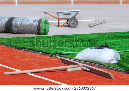 Laying artificial turf on the sports field of the stadium. Green grass carpet rolls and orange soft crumb rubber tracks. Tools and materials on a construction site without people. Sunny day.
