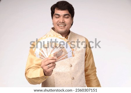 Portraits of Smiling Handsome Indian Man showing indian rupees. Wearing traditional ethnic kurta with dark red jacket in shopping festival. celebration theme. studio background 