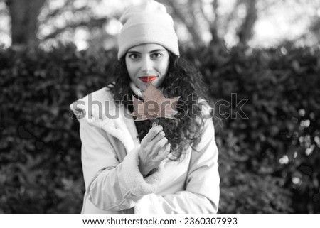pretty young woman with curly brown hair wears a woollen hat for the cold and covers her face with a dry leaf from a tree. Autumn and cold concept. Monochrome photo