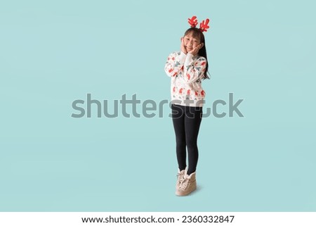 Cheerful young Asian girl wearing a Christmas sweater with reindeer horns, Happy smiling hand holding face standing posing full body portrait, isolated on pastel plain light blue background.