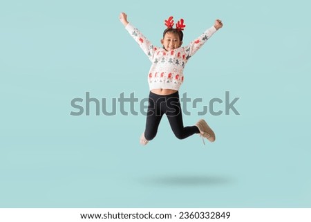 Cheerful young Asian girl wearing a Christmas sweater with reindeer horns, Happy smiling dance jumping have fun full body portrait, isolated on pastel plain light blue background.