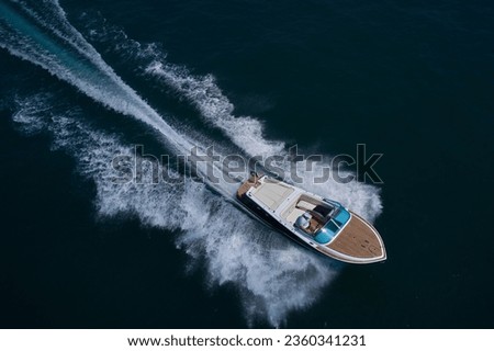 Luxury wooden big speedboat fast moving on dark water top view. Boat movement on the water. Motor boat in motion. Expensive wooden big boat with people moving on the water aerial view. Royalty-Free Stock Photo #2360341231
