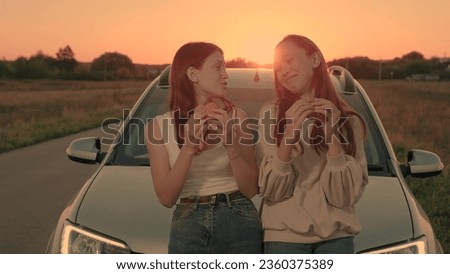 Positive women rest and eat burgers sitting on car bonnet at back sunset light. Happy women with fresh sandwiches relax during car trip. Young women with hamburgers lean on car bonnet in field Royalty-Free Stock Photo #2360375389