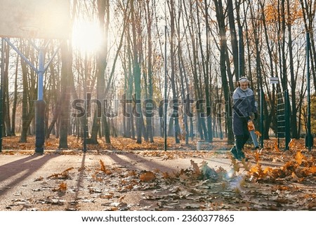 Focused male janitor cleaning outdoors baskeball court from leaves with hand blower. Side view of senior man in uniform taking care of city area during autumn time. Work, fall season concept.