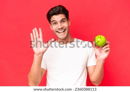 Young caucasian man with an apple isolated on red background saluting with hand with happy expression
