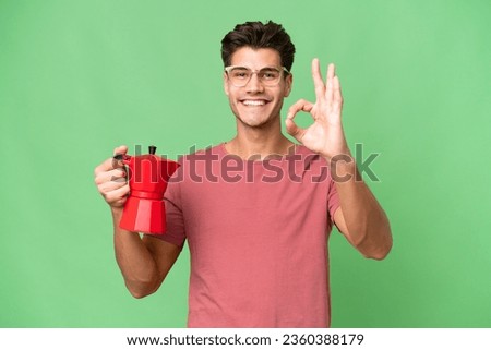 Young caucasian man holding coffee pot over isolated background showing ok sign with fingers