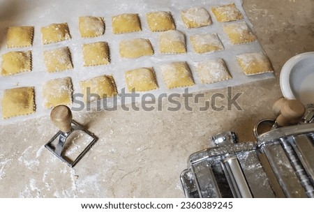 Homemade ravioli, floured and ready to be cooked, pictured with pasta making machine and ravioli press. Royalty-Free Stock Photo #2360389245