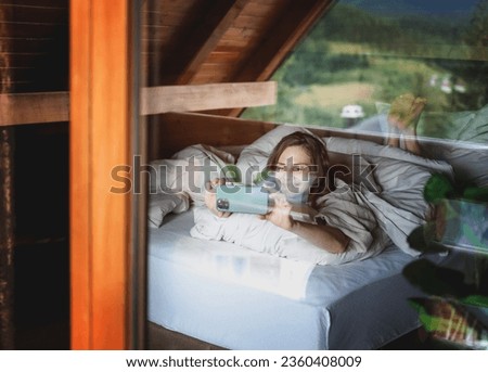 Young relaxed cheerful woman lying in bed enjoying the morning in a village house in the forest taking photos on a smartphone