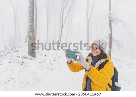 Young caucasian woman in a yellow jacket and hat walking in a snowy forest taking pictures of nature on the phone