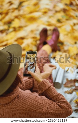 Rear view of fashionable woman in hat taking photo on camera phone while relaxing in beautiful autumn park. Focus on picture in smartphone