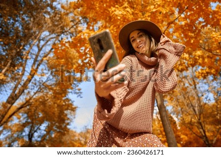 Autumnal style trendy shot. Stylish woman in hat and knit sweater captures the moment with selfie on smartphone at park with orange leaves