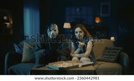 Man horror maniac and young girl sitting on the sofa in the room, girl eats pizza, both looking at the camera.