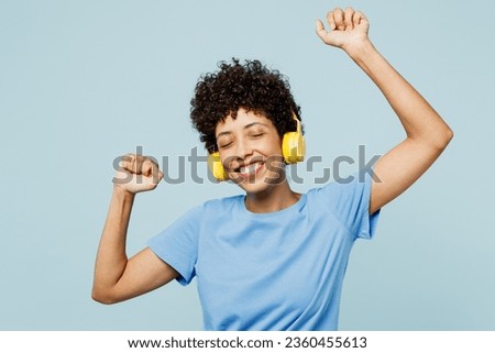 Young happy woman of African American ethnicity wear t-shirt casual clothes listen to music in headphones raise up hands dance isolated on plain pastel light blue cyan background. Lifestyle concept