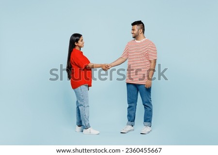 Full body young couple two friends family Indian man woman wear red clothes t-shirts together hold hands folded handshake gesture isolated on plain blue background. Friendship business greet concept Royalty-Free Stock Photo #2360455667