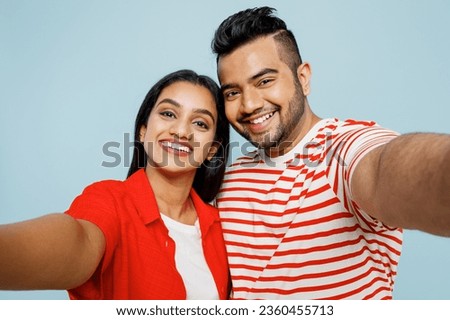 Close up young couple two friends family Indian man woman wear red casual clothes t-shirts together doing selfie shot pov on mobile cell phone hug isolated on pastel plain blue cyan color background
