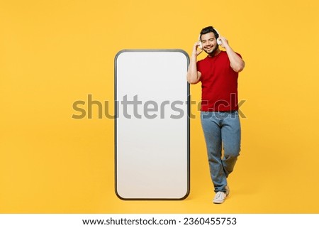 Full body fun young Indian man wears red t-shirt casual clothes big huge blank screen mobile cell phone smartphone with area listen oto music in headphones isolated on plain yellow orange background