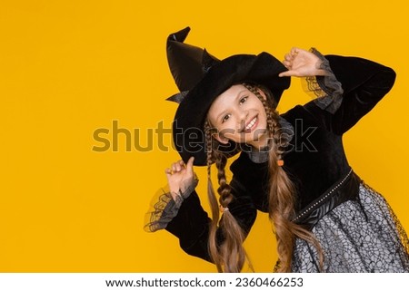 A young witch in a black fluffy dress and long pigtails supports her pointed black hat with her hands. portrait of an attractive witch on a yellow isolated background.