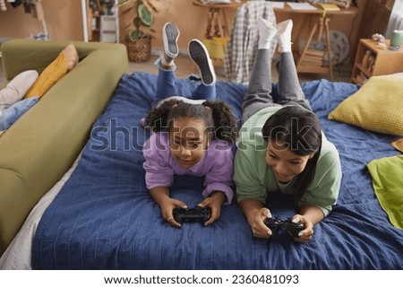 High angle portrait of happy black mother and daughter playing video games together lying on bed at home