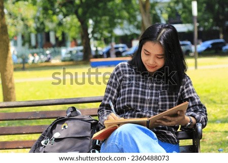 smiling asian beautiful young woman enjoy writing to do list and idea in notebook in outdoors city park