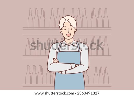 Man works in winery standing with crossed arms near racks filled with bottles wine. Guy in apron poses in warehouse with wine, taking care of compliance with storage conditions for expensive alcohol. Royalty-Free Stock Photo #2360491327