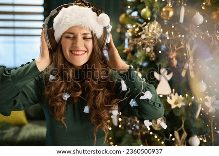 Christmas time. happy trendy housewife with Santa hat in green dress listening to the music with headphones near Christmas tree in the modern living room. Royalty-Free Stock Photo #2360500937