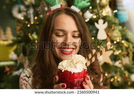 Christmas time. smiling stylish 40 years old housewife with festive hot chocolate cocktail in traditional Christmas sweater near Christmas tree at modern home. Royalty-Free Stock Photo #2360500941