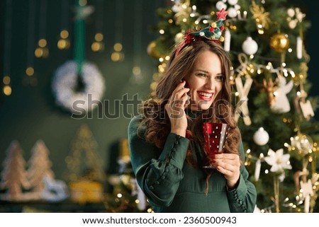 Christmas time. happy modern woman with glass in green dress using a smartphone near Christmas tree in the modern living room. Royalty-Free Stock Photo #2360500943