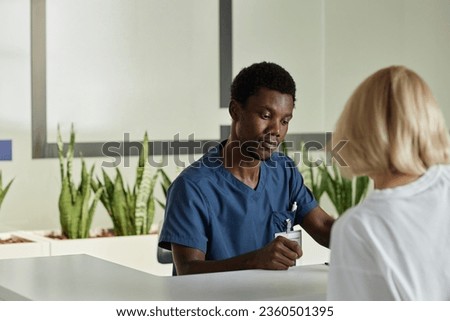 Young African American male clinician looking at female patient signing medical document while standing by reception counter in front of him Royalty-Free Stock Photo #2360501395