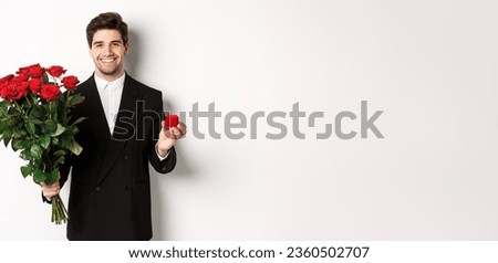 Image of handsome man in black suit, holding bouquet of red roses and a ring, making a proposal, smiling confident, standing against white background.