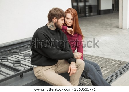 Stylish beautiful fashionable young couple, handsome hipster man and a redhead pretty girl in a fashionable hoodie are sitting together on the street