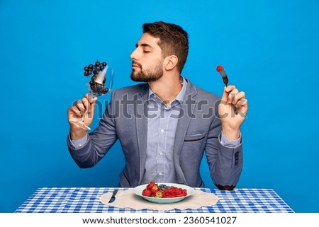 Businessman eating delicious berries, strawberries, grapes and blueberries against blue background. Concept of food, creativity, summer, taste, health. Pop art photography. Copy space for ad