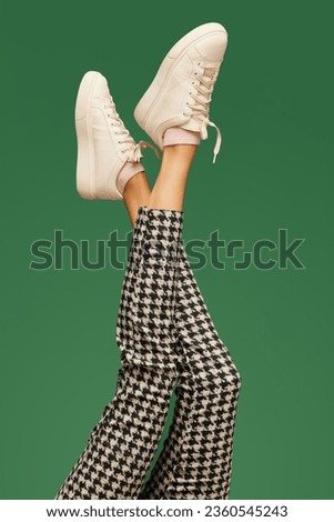 Female legs in white casual sneakers and in checkered pants over green background. Smart casual, business style. Colorful photography. Concept of fashion, creativity, imagination. Copy space for ad