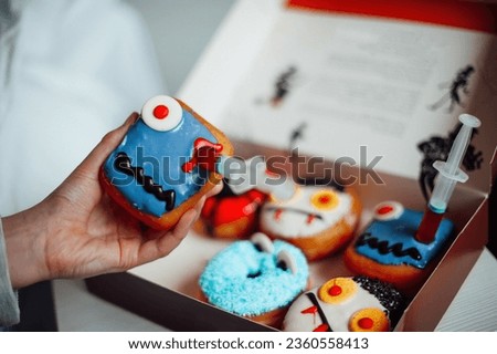 Donuts with Halloween decoration of monsters with spooky face and zombie by syringe with sugar syrup. Different colorful traditional sweets and doughnuts of all hallows eve at autumn season indoor