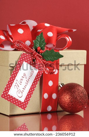 Natural modern trend Christmas gift wrapping with brown kraft gift box and red and white polka dot ribbon with holly and gift tag on dark red background. Vertical.