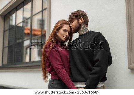 Beautiful fashion couple of lovers in stylish clothes with a pullover are standing on the street. Handsome hipster man in a black sweatshirt and a beautiful redhead woman in a red hoodie hug outdoors