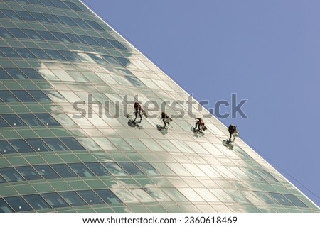 Team of men cleaning windows of a high rise glass skyscraper; professionals maintaining a modern office block; window cleaners abseiling or rappelling down a glass skyscraper
